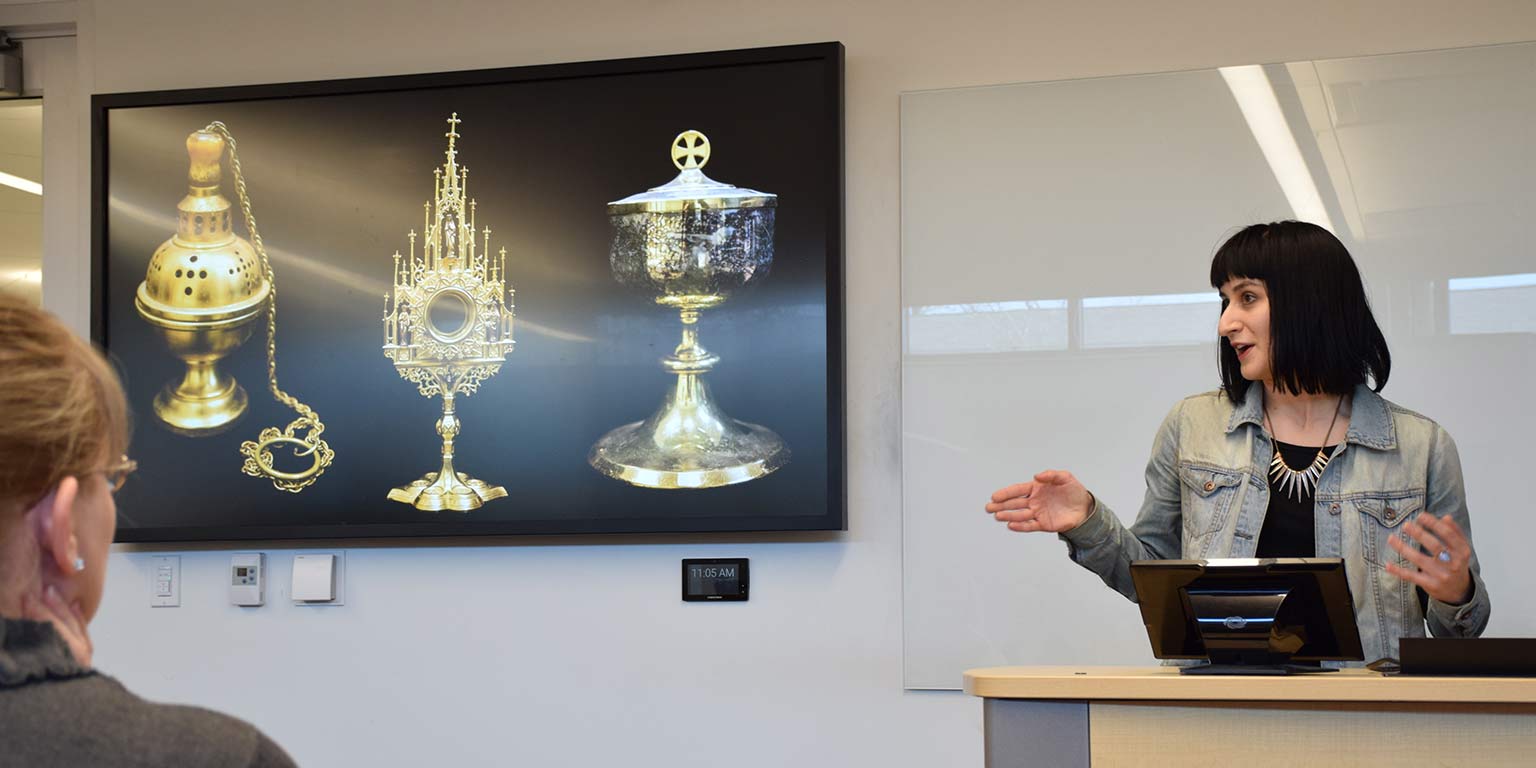 A woman with short black hair stands behind a lectern, gesturing towards a powerpoint slide that shows gold artifacts.  