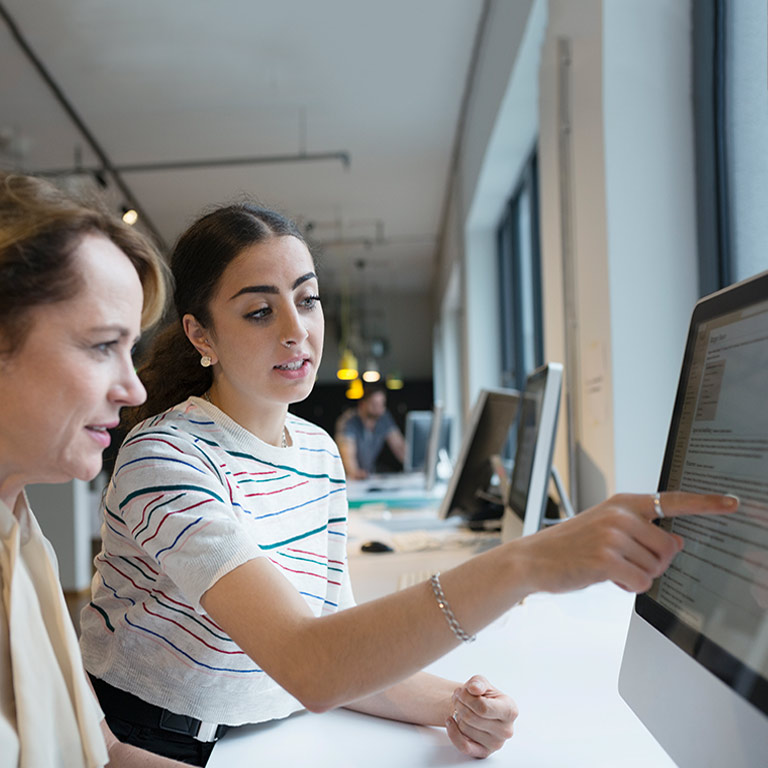 Two women sit side by side in front a computer; one points to the screen in front of them.