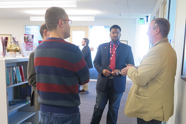 Four men stand in a circle discussing. They are wearing lanyards around their necks. 