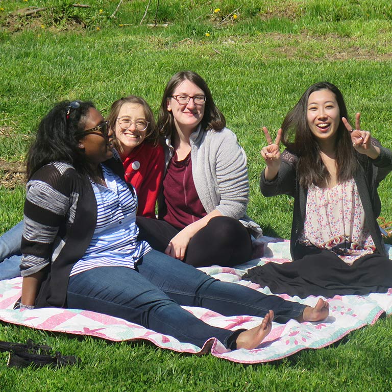 Four female students sit on a green lawn laughing; one is flashing a peace sign with her fingers to the camera. 