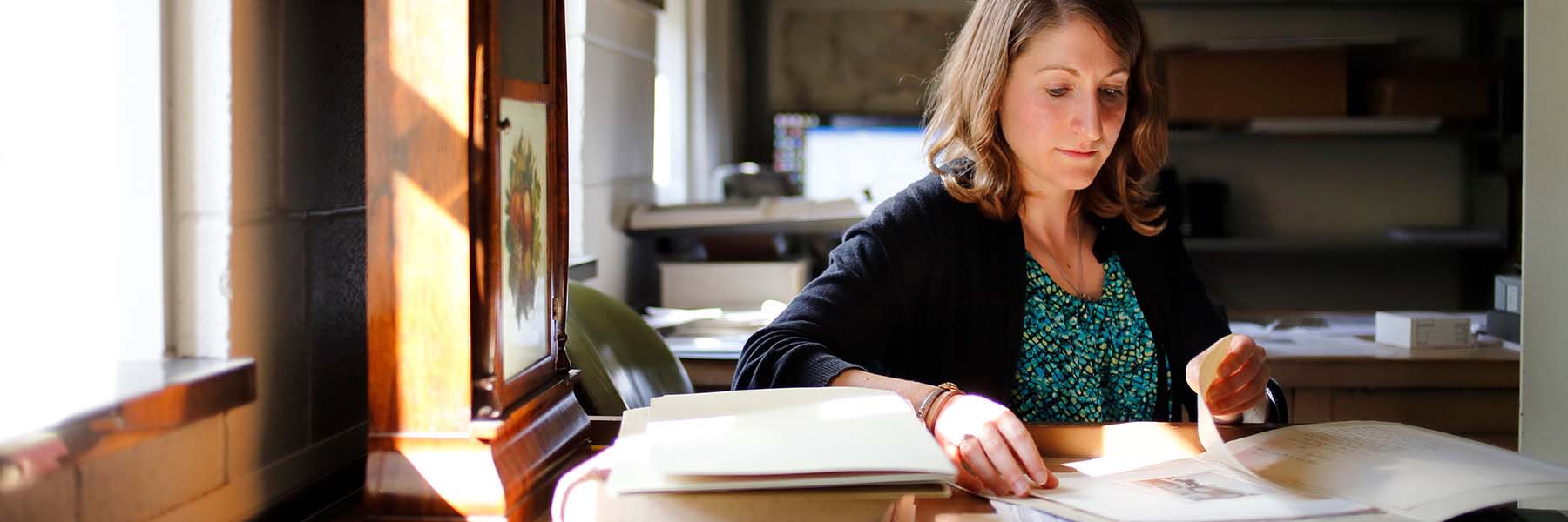 A female student sits at a library desk reviewing an open book.