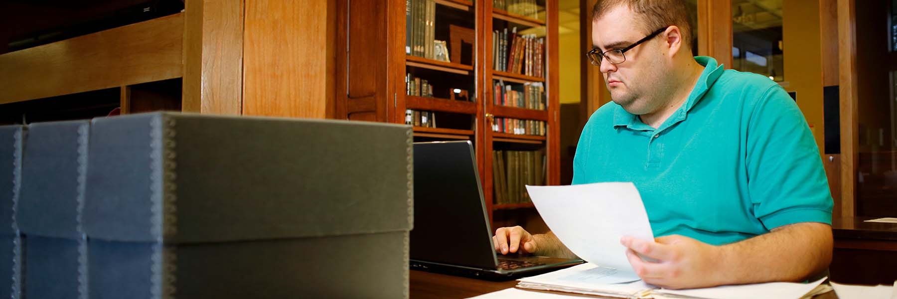 A male student sits at a library table taking notes while examining an open book. 