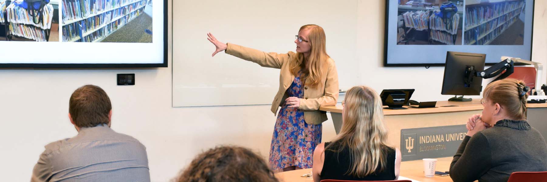 An instructor stands at the front of a classroom, gesturing to a Powerpoint slide that shows a shelf of books.