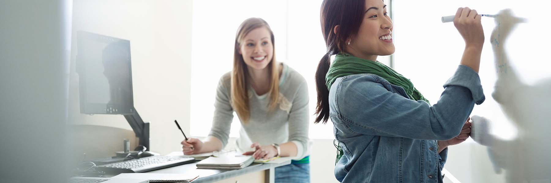 One female students writes on a classroom whiteboard, while a student behind her takes notes in a personal notebook. 