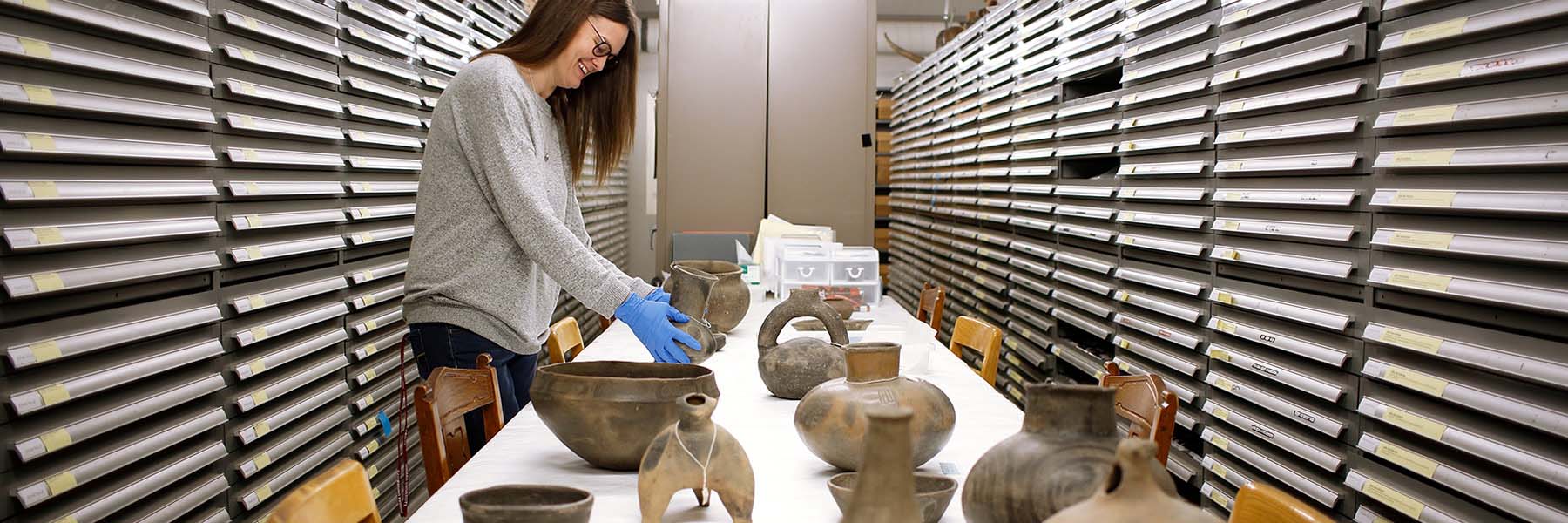 A student wearing blue latex gloves places a piece of ancient pottery on a table with other ancient pottery. 