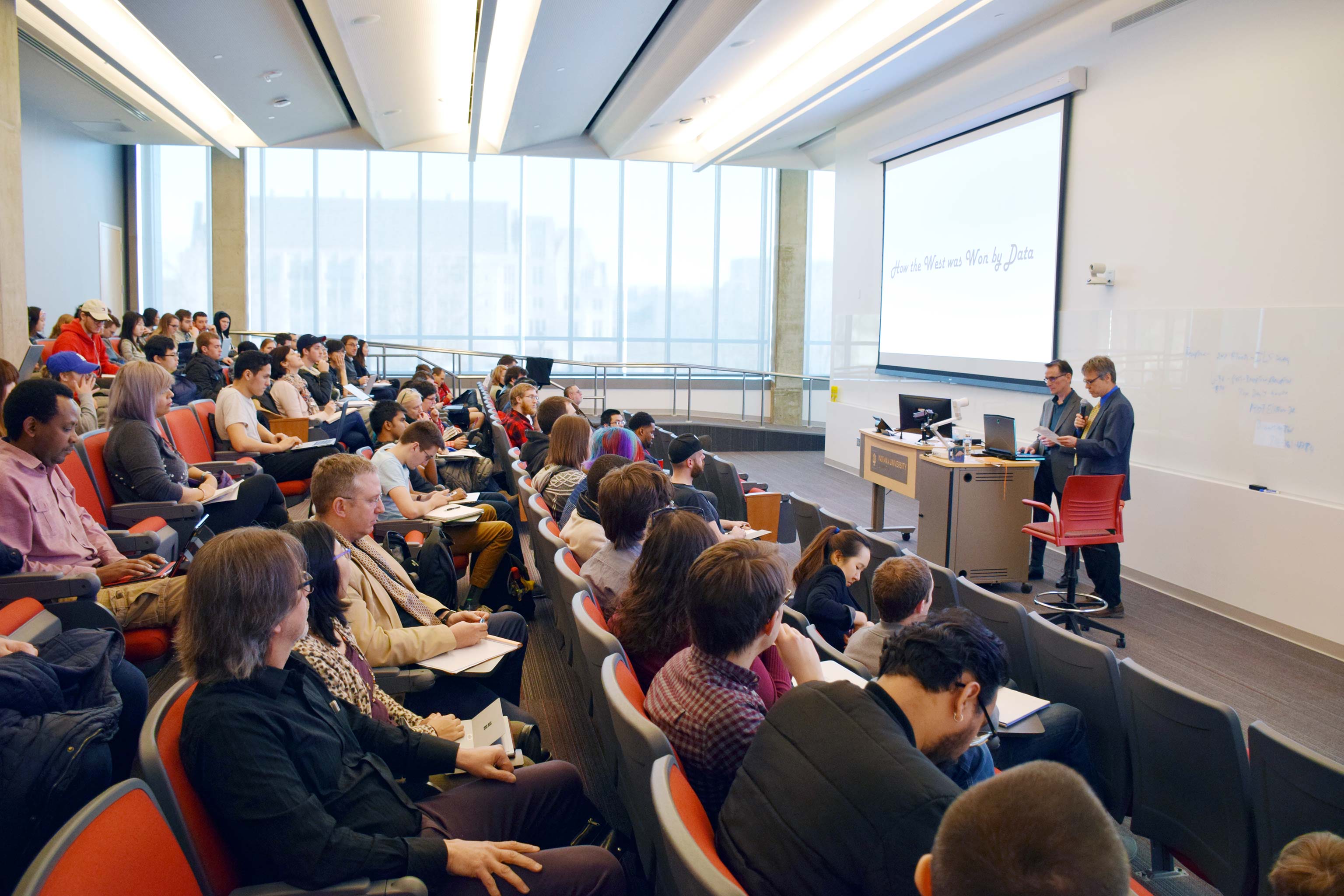 Two presenters stand behind a lectern in front of a full lecture hall. 